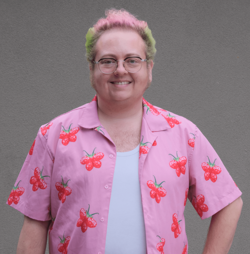 A photo of Tracey Simmons. His hair is green and pink, and his button up shirt is pink with strawberries on it. 